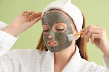 Wall Mural - Young woman with applied facial mask and brush on green background, closeup