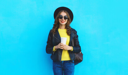 Wall Mural - Stylish happy young woman holding cup of coffee in black hat, leather jacket on blue background