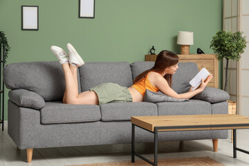 Beautiful young woman reading book on cozy sofa in living room