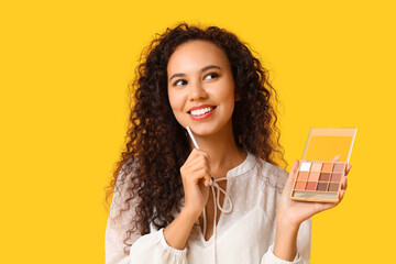 Wall Mural - Beautiful young happy African-American woman with brush and eyeshadows palette on yellow background