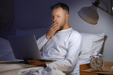 Wall Mural - Young man with laptop suffering from insomnia on bed in bedroom at late night