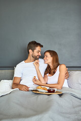 Poster - Couple, eating and smile in bed for breakfast at hotel with room service for love and valentines day. Man, woman and happy in morning in bedroom for support, food and celebration together on vacation