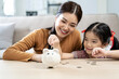 © NAMPIX - Financial education. asian family, mother and daughter with pig piggy bank counting savings at home on table.