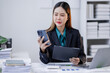 © David - Asian woman sit at workplace desk holds cellphone staring at laptop, synchronize data between computer and gadget in office, use corporate devices and business application, plan work, use organizer