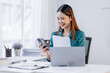 © David - Asian woman sit at workplace desk holds cellphone staring at laptop, synchronize data between computer and gadget in office, use corporate devices and business application, plan work, use organizer