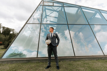 A man in a suit stands in front of a large glass building, holding a bouquet of flowers. Concept of elegance and sophistication, as the man is dressed in a formal attire