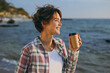 © ViDi Studio - Close up young woman wear shirt casual clothes hold takeaway delivery craft paper brown cup coffee to go rest on sea ocean sand shore beach outdoor seaside in summer day free time. Lifestyle concept.
