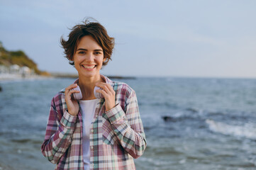 Wall Mural - Young smiling happy woman wear shirt casual clothes hold headphones on neck listen to music look camera rest on sea ocean sand shore beach outdoor seaside in summer day free time. Lifestyle concept.