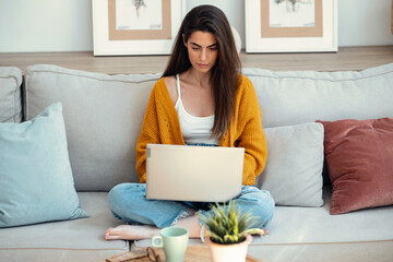 Wall Mural - Beautiful young woman working with her laptop while sitting on a couch at home.