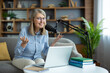 © Liubomir - Smiling senior woman recording podcast, sitting on sofa with laptop and microphone. Engaged in online communication, sharing knowledge, and connecting with audience. Concept of remote work and