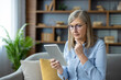 © Liubomir - Senior woman with glasses reading on tablet in modern living room thoughtfully. She seems deeply engaged with information on the device showing concentration and curiosity.