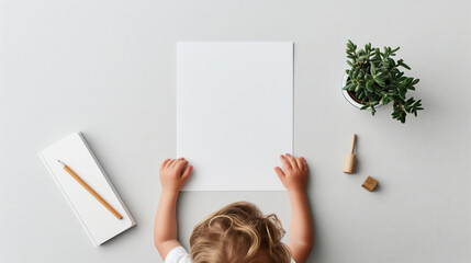 Wall Mural - Top view of a child holding a blank sheet of paper, with a notebook and pencil on the left, a wooden toy on the right, and a potted plant on a light grey background.