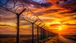 © Wanlop - Foreboding state border with high razor-sharp barbed wire fencing stretches across the horizon, set against a vibrant orange and pink sunset backdrop.