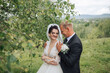 © Vasil - A bride and groom are standing in a field with trees in the background. The bride is wearing a white dress and the groom is wearing a suit. They are both smiling and seem happy