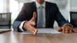 © Iana - Businessman extending his hand for a handshake at an office desk with a contract document on the table