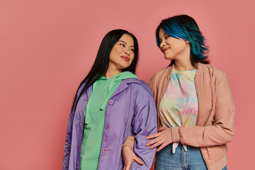 Asian mother and teenage daughter in casual attire stand gracefully in front of a vibrant pink studio wall.