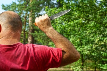 one dangerous kitchen sharp shiny iron dirty knife with a wooden handle in the hand near the head of an aggressive bald man in a red T-shirt near green trees during the day on the street