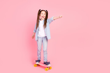 Poster - Full size photo of small girl dressed denim shirt pants riding skateboard to empty space raising fist isolated on pink color background