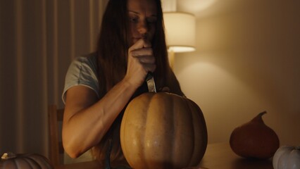 Wall Mural - In the Dark by Warm Lamp and Candlelight on the Table, a Woman Carving a Jack-o'-Lantern from a Pumpkin. She Thrusts the Knife Downward. Slow Motion.