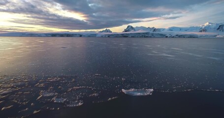 Wall Mural - Low angle flight over polar icy ocean in Antarctica. Snow-covered mountains range under colorful sunset clouds sky. Tranquil scene icy landscape, wild nature background. South Pole travel, exploration