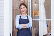 © Ekkasit A Siam - Asian spa owner smiling at entrance of spa. Wearing a blue apron and light blue shirt, arms crossed. Open sign on door, welcoming clients to a clean, inviting, professional spa environment.