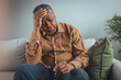 © Dragana Gordic - Elderly Black Man Experiencing Discomfort on Couch. A senior black male grips his glasses, showing signs of distress while seated alone on a sofa, reflecting potential health concerns