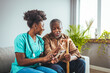 © Dragana Gordic - A mid-adult Black nurse in scrubs attentively assists an elderly Black man with his medication, highlighting compassionate in-home care in a cozy living room setting.