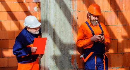 Two builders in hard hat on construction site. Male builder in uniform with smartphone during work break. Foreman or inspector with clipboard checklist checks work of construction crew at workplace.