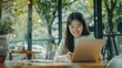 © vefimov - A young woman in a white blouse smiling at her laptop, working in a contemporary coffee shop with natural light and plants.