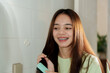 © Ekkasit A Siam - Young Asian girl brushing long straight brown hair in bathroom. Wearing light green ribbed top. Indoor setting with tiled walls. Expressing joy and self-care in morning routine.
