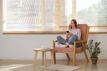 Poster - Young woman using mobile phone while sitting in armchair with cushion in room