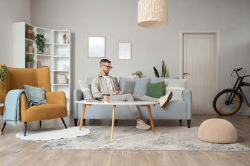 Wall Mural - Young bearded man in eyeglasses using laptop on sofa at home