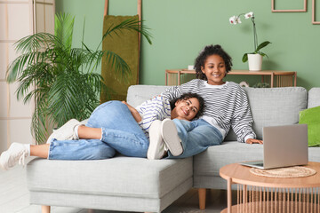 Sticker - Beautiful young happy African-American mother and her daughter with laptop watching movie on sofa at home