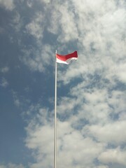 The red and white Indonesian flag flies high on a pole against the sky in the background. sunny, cloudy