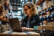 © vefimov - Businesswoman working on laptop in a warehouse setting, engaging with technology for business operations.