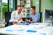 © Phushutter - An Italian male and a Caucasian male sit at business desk in office meeting room, discussing financial terms, investment strategies, managing funds with computer monitor displaying relevant data.