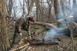© soleg - Two men make a campfire for cooking in the forest, father and son sitting on a log while hiking and outdoor activities, early spring landscape