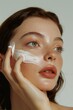 © Vadym - Close-up of a woman face applying white cream. Her left hand holds the cream while her right hand rests on her cheek. The background is gray and simple, contrasting with her vibrant skin and hair.