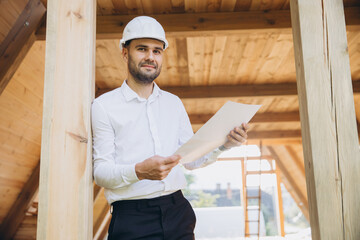 Architect wooden buildings. Man stands under frame wooden house. Man architect studies documentation or blueprints. Guy in protective helmet at construction site. Architect under timber framed house