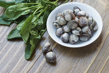 Canvas Print - Close-up of stacked raw cockles on white dish with spinach leaves on wood floor, South Korea
