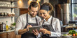 © Anhen Design - Two restaurant workers, wearing aprons, stand in the kitchen using a touchscreen tablet. They look focused and engaged in their task, with shelves of ingredients and kitchenware behind them
