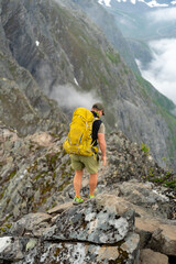 male backpacking hiker with yellow backpack climbing Romsdalseggen trail in Andalsnes Norway in summer with rocky mountains in the background