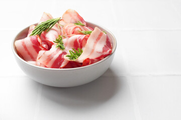 Wall Mural - Slices of raw bacon and rosemary in bowl on white tiled table, space for text