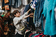 © qunica.com - A young individual browsing clothes in a thrift store, exploring second-hand fashion and sustainable shopping options.