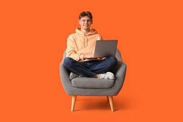 Canvas Print - Handsome young man with modern laptop sitting in armchair on orange background