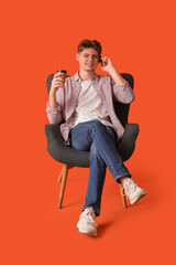 Poster - Handsome young man with paper cup of coffee talking by mobile phone and sitting in armchair on orange background