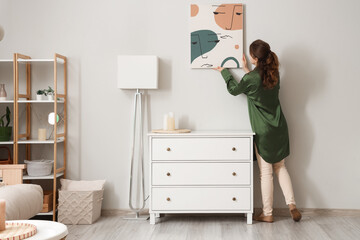 Poster - Young woman hanging picture near chest of drawers in stylish room