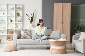 Wall Mural - Young African-American woman in headphones using laptop on sofa at home