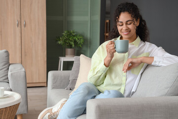 Sticker - Young African-American woman with cup of tea resting on sofa at home