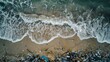 © HanThar Fusion - aerial view of the ocean waves crashing onto an overcast beach, with scattered plastic bottles and other debris on its shore